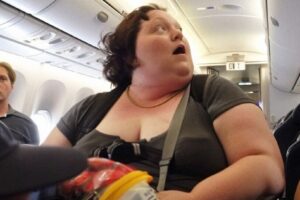 The rich man makes fun of the poor, obese woman on the plane when he hears the captain talking to her.